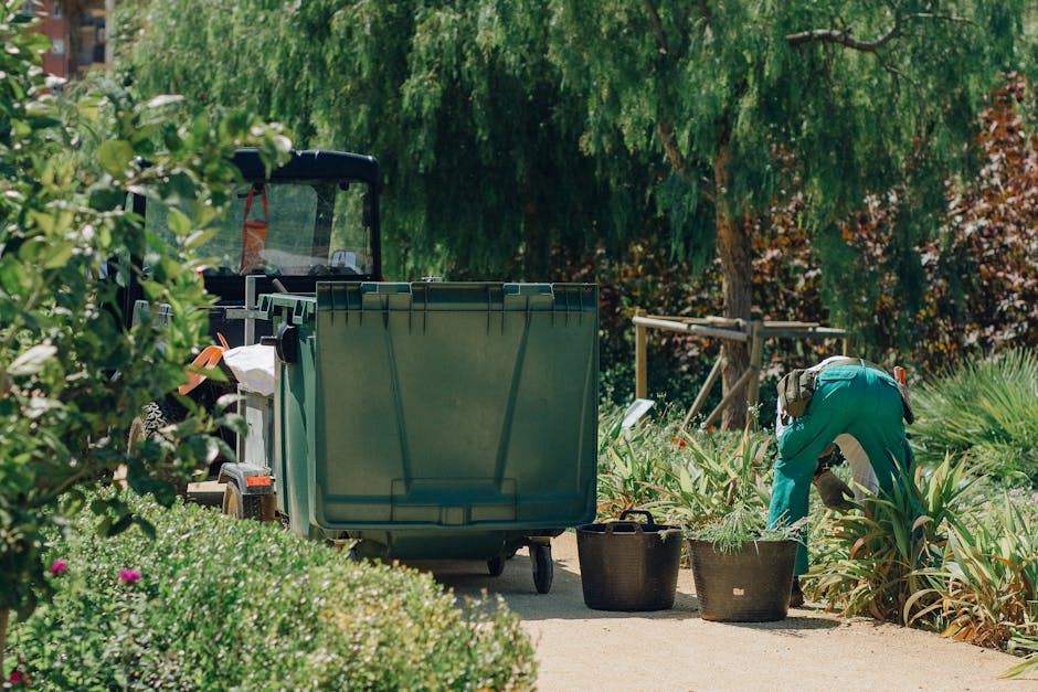 A gardener wearing teal overalls is kneeling beside a landscaped garden bed, tending to plants with a gardening tool. The garden features a variety of lush, green foliage, including tall, spiky plants