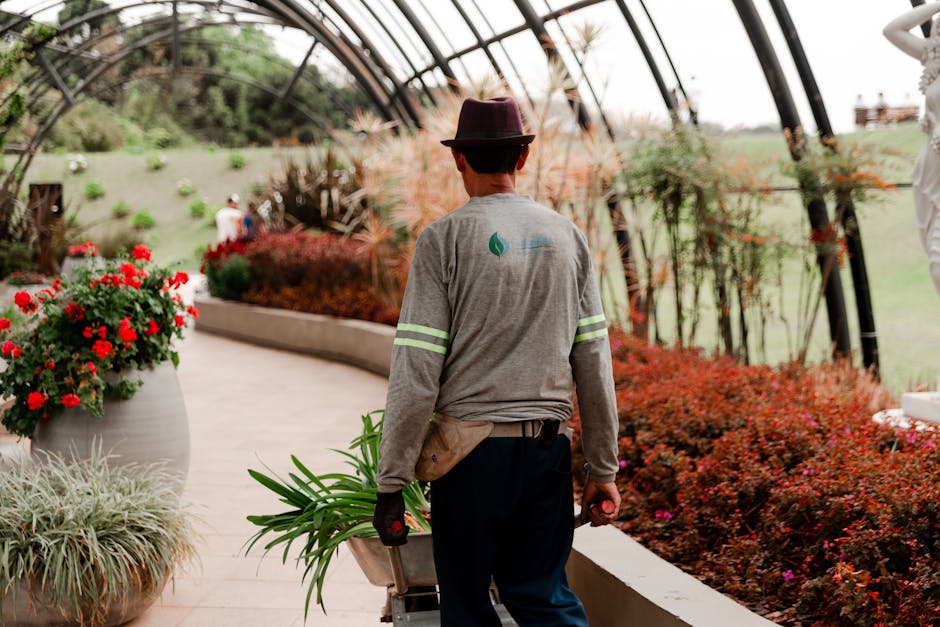A gardener wearing teal overalls is kneeling beside a landscaped garden bed, tending to plants with a gardening tool. The garden features a variety of lush, green foliage, including tall, spiky plants